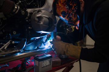 Worker in helmet with flames welding the steel part of a motorbike using an argon welding machine and gloves in a garage