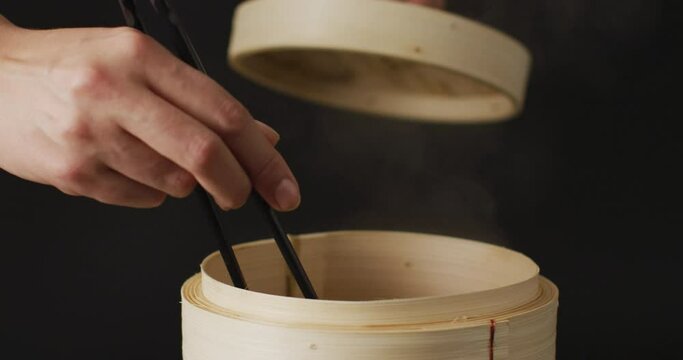 Hand holding chopsticks with gyoza dumpling in bamboo steamer on black background