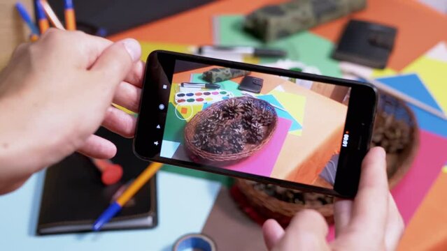 Hands Taking Photos of a Creative Workplace Desk on Smartphone. Palette of Watercolor, Colored Pencils, Cardboard Scattered on Table. Art materials. Concept of school, lessons, education, creativity.