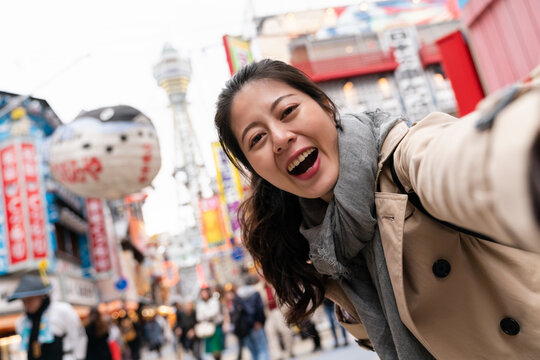 self portrait of happy Asian Japanese female tourist looking at camera while taking picture with tsutenkaku tower at background in nostalgic Shinsekai shopping area of Osaka Japan