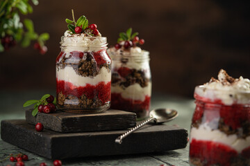 Latvian, scandinavian traditional rye whole grain bread layered dessert with whipped cream and cowberry jam served in glass jars on brown wooden board, table with berries, copy space.