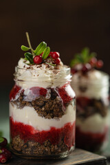 Latvian, scandinavian traditional rye whole grain bread layered dessert with whipped cream and cowberry jam served in glass jars on brown wooden board, table with berries, copy space.