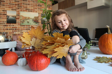 A little girl is sitting among pumpkins with a bouquet of dried maple leaves in her hands, preparing for the Halloween holiday, home and street decor.