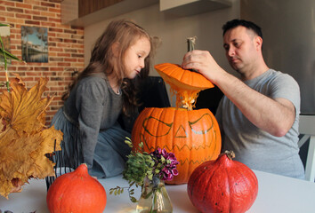 A little girl with long hair and her dad are preparing Jack's Lantern from a large orange pumpkin. The tradition of preparing for the Halloween holiday. Decor of dried leaves, flowers and candles 