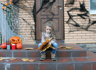 A sad girl with big eyes is sitting on the porch against the background of a house with a Halloween decoration holding a dry maple leaf. Traditions of decorating houses with cobwebs, pumpkins, spiders