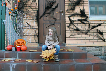 A sad girl with big eyes is sitting on the porch against the background of a house with a Halloween decoration holding a dry maple leaf. Traditions of decorating houses with cobwebs, pumpkins, spiders