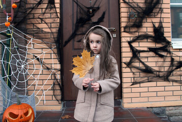 A girl with big eyes in warm clothes on the background of a house with a Halloween decoration holds a dry maple leaf. Traditions of decorating houses with cobwebs, pumpkins, spiders and dry leaves