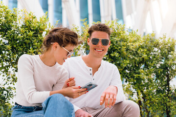 Young modern couple use smartphone sitting bench outdoor in office district.