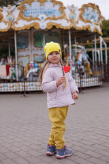 Little girl eating red heart lollipop in child attraction park, autumn clothes. Yellow hat and pink jacket. Child licking sweets. Funny and happy child having fun outdoors.