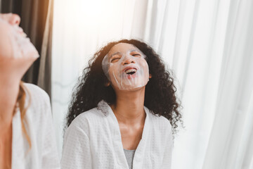 Beautiful young mixed race African American woman in white bathrobe applying a revitalising mask her friend's face. Young happy girls doing facial skin beauty treatment with friends at home.