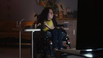 Fototapeta na wymiar Woman with disability in a modern motorized wheelchair driving into living room and watching TV series in the evening while having dinner