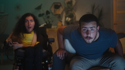 Shocked woman with physical disability in a wheelchair and man wondering and looking at each other, while watching TV show together at home in the evening