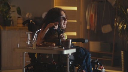Naklejka premium Cheerful woman with physical disability in a wheelchair playing video game on TV using gamepad while being alone at home in the evening