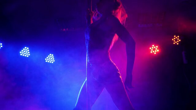 Silhouette of a slender young woman in lingerie in a nightclub near a pole, she dances in the light of multi-colored spotlights. Pole dance.