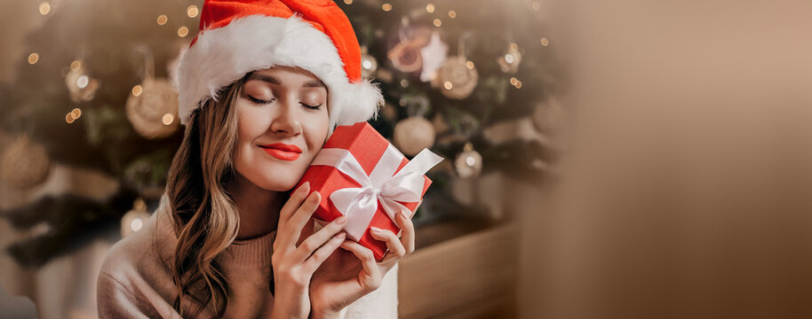 Christmas gifts concept. A young Caucasian girl hugs a red gift box and makes a wish on New Year's Eve and dreams against the background of a New Year tree and a garland. Copy space
