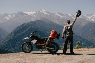 Male biker traveler standing in mountains near his motorcycle holding helmet in raised hand