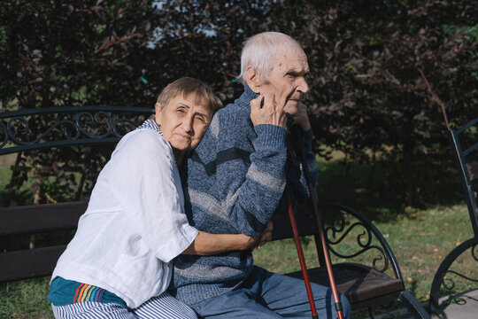 Elderly man with trekking sticks and woman couple, husband and wife sitting on park bench hugging and enjoying sunny day