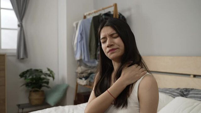 Asian girl having a crick in the neck is exercising her stiff muscles and massaging her painful shoulders after getting up in the morning in the bedroom at home