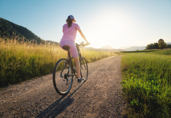Woman is riding a mountain bike on gravel road at sunset in summer. Colorful landscape with sporty...