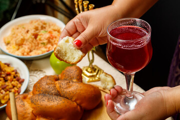 A glass of wine and a piece of challah in women's hands kiddush on the day of Rosh Hashanah in women's hands over a festive table with a menorah.
