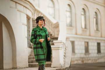 Fototapeta na wymiar Outdoor fashion portrait of an elegant fashionable brunette woman, model in a stylish cap, green dress, posing at sunset in a European city in autumn.