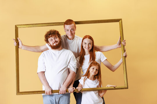 Joyful redhead family looking through picture wooden frame together. Young men, woman and kid spend time together at studio photo shoot.