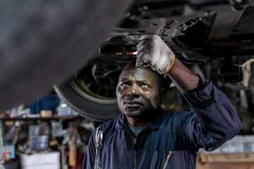 Obraz na płótnie Canvas African auto mechanic fixing underneath vehicle in auto repair service shop, black man working in car garage , Car maintenance concept