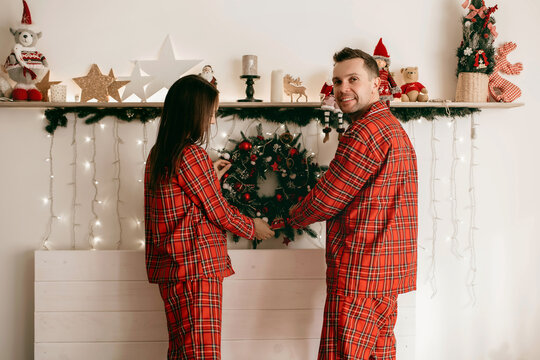 A Man And A Woman Dressed In Red Pajamas Decorate Their Bedroom With Christmas Decor. A Loving Couple Is Preparing For The Christmas Holidays And Decorating Their Home Together