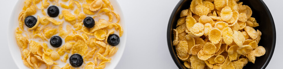 top view of bowls with dry and wet corn flakes in milk with blueberries isolated on white, banner.