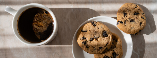 top view of cup of black coffee near white saucer with chocolate chip cookies on marble surface, banner.