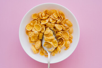 top view of bowl with corn flakes, milk and spoon on pink background.