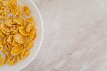 top view of crispy corn flakes in bowl with spoon and milk on marble surface.