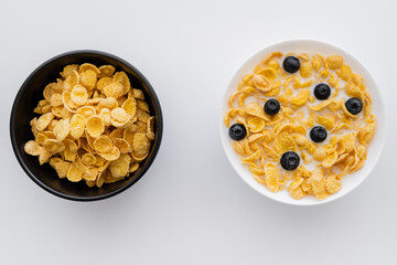 top view of bowls with dry and wet corn flakes in milk with blueberries isolated on white.