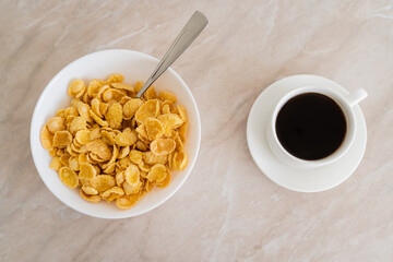 top view of bowl with corn flakes and spoon near cup of coffee on marble surface.