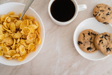top view of bowl with corn flakes near chocolate chip cookies and cup of coffee on marble surface.