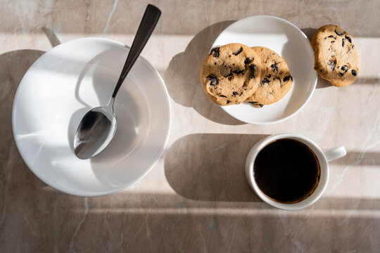 top view of cup of black coffee near chocolate chip cookies and saucer with spoon on marble surface.