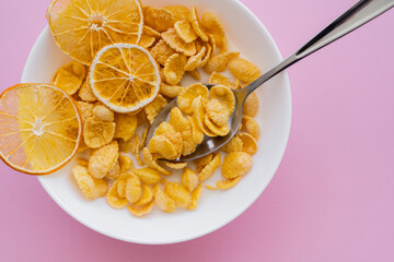 top view of dried oranges in bowl with corn flakes and spoon on pink background.