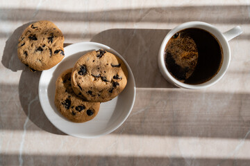 top view of cup of black coffee near white saucer with chocolate chip cookies on marble surface.