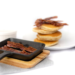 Closeup of mini cast-iron skillet with crispy bacon and pancake stack in the background isolated against white background