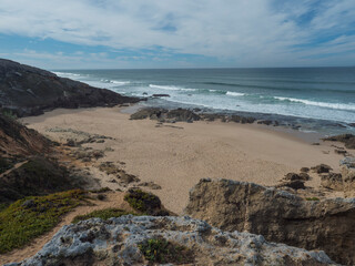 View of empty small sand beach with ocean waves and sharp rock and cllifs at wild Rota Vicentina coast near Porto Covo, Portugal.