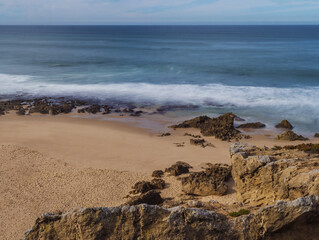 View of empty small sand beach with with long exposure blurred ocean waves and sharp rock and cllifs at wild Rota Vicentina coast near Porto Covo, Portugal.