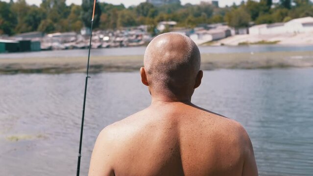 Angler Standing on Riverbank, Catching Fish, Looking into the Distance on Sunset. Back view of a bald man enjoying a fishing trip. Leisure, hobby, lifestyle. Nature, summer landscape. Weekend.