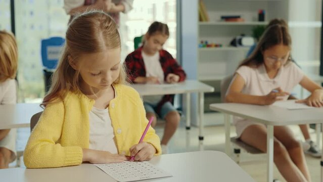Exam in elementary school. Beautiful blonde pupil sits at a first desk in the classroom and fills out tests thinks about answers, and the teacher standing in the background.
