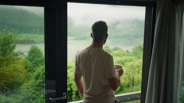 Man standing at the window, enjoying view at nature, drinking coffee