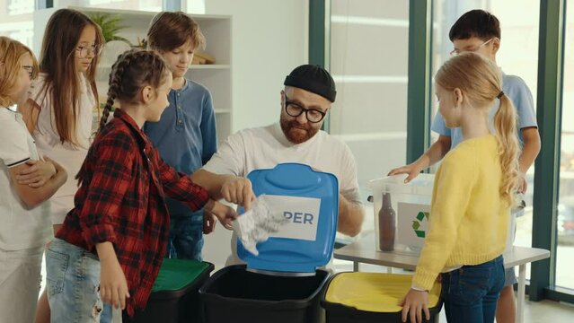 Male teacher teach where to sort the rubbish. Garbage sorting class, teacher tells and asking children were they may put the glass bottle and the plastic cup the pupils answer right.