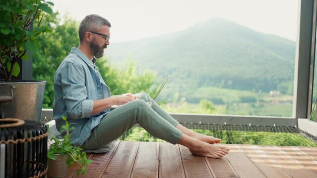 Mature man working on laptop, on terrace of tiny house, enjoying the nature view.