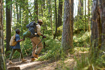 Side view of cute schoolboy with rucksack and trekking sticks following his grandfather with...