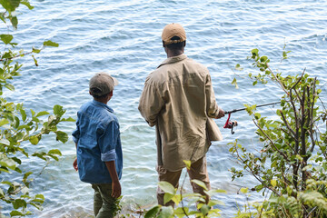 Rear view of senior black man with rod and his grandson standing in front of lake or river and...