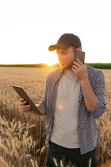 Bearded farmer talking on mobile phone and looking at digital tablet in agricultural field at sunset