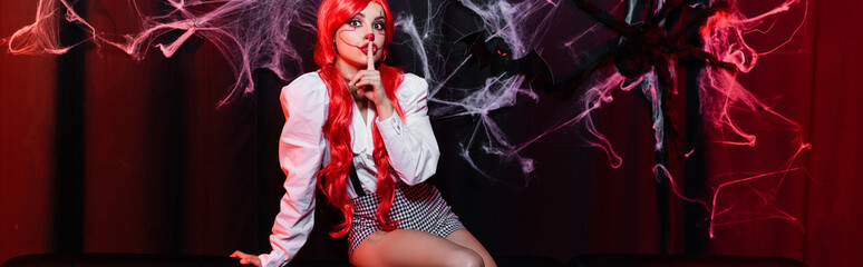 young redhead woman in halloween makeup showing hush sign on dark background with spiderweb, banner.